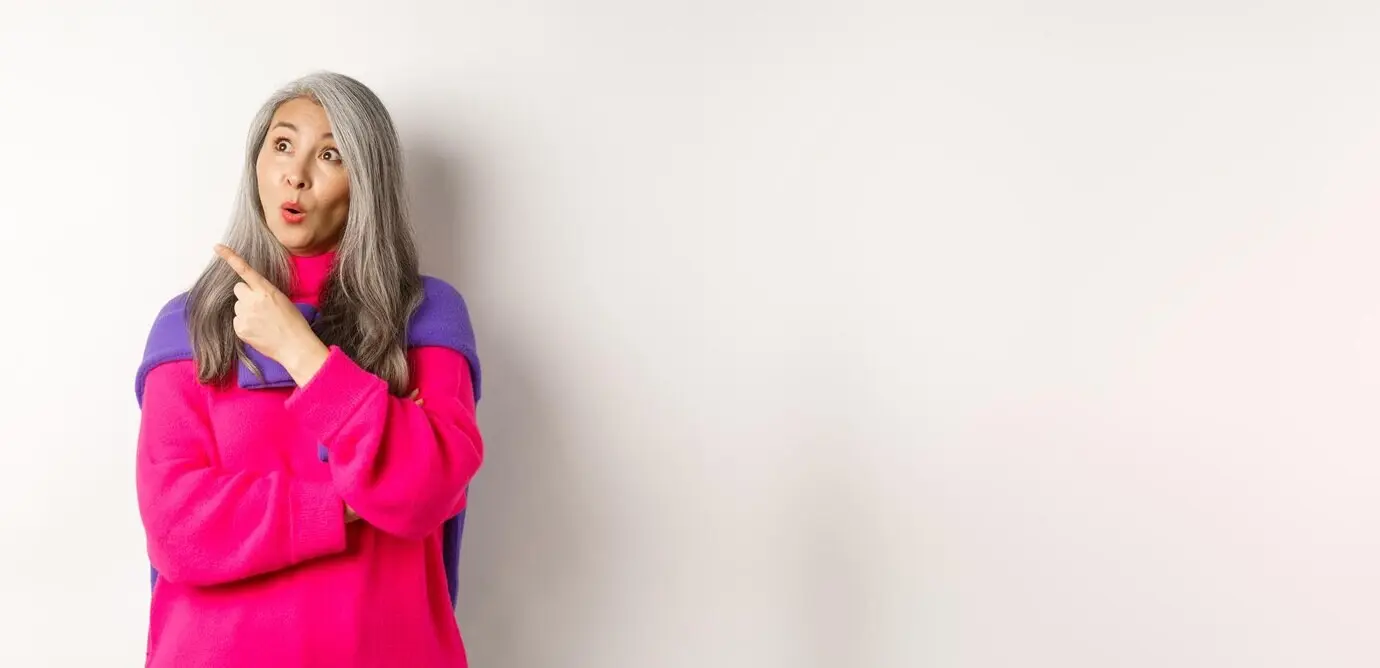 Заинтересованная пожилая азиатка изучает промоакцию, смотрит и указывает пальцем влево, стоя на белом фоне.