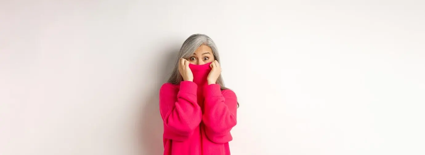 Портрет смешной азиатской бабушки, прячущей лицо в воротнике свитера, дурашливо заглядывающей в камеру и стоящей.